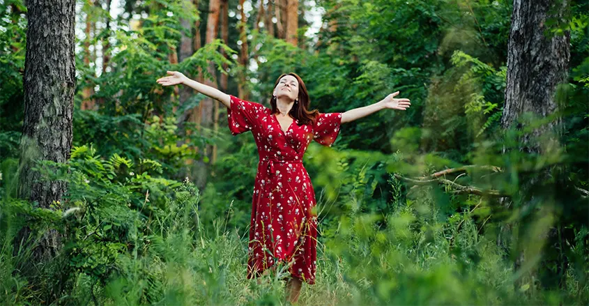 Foto de mujer en el bosque rodeada de árboles con los brazos en cruz reconectando con la naturaleza